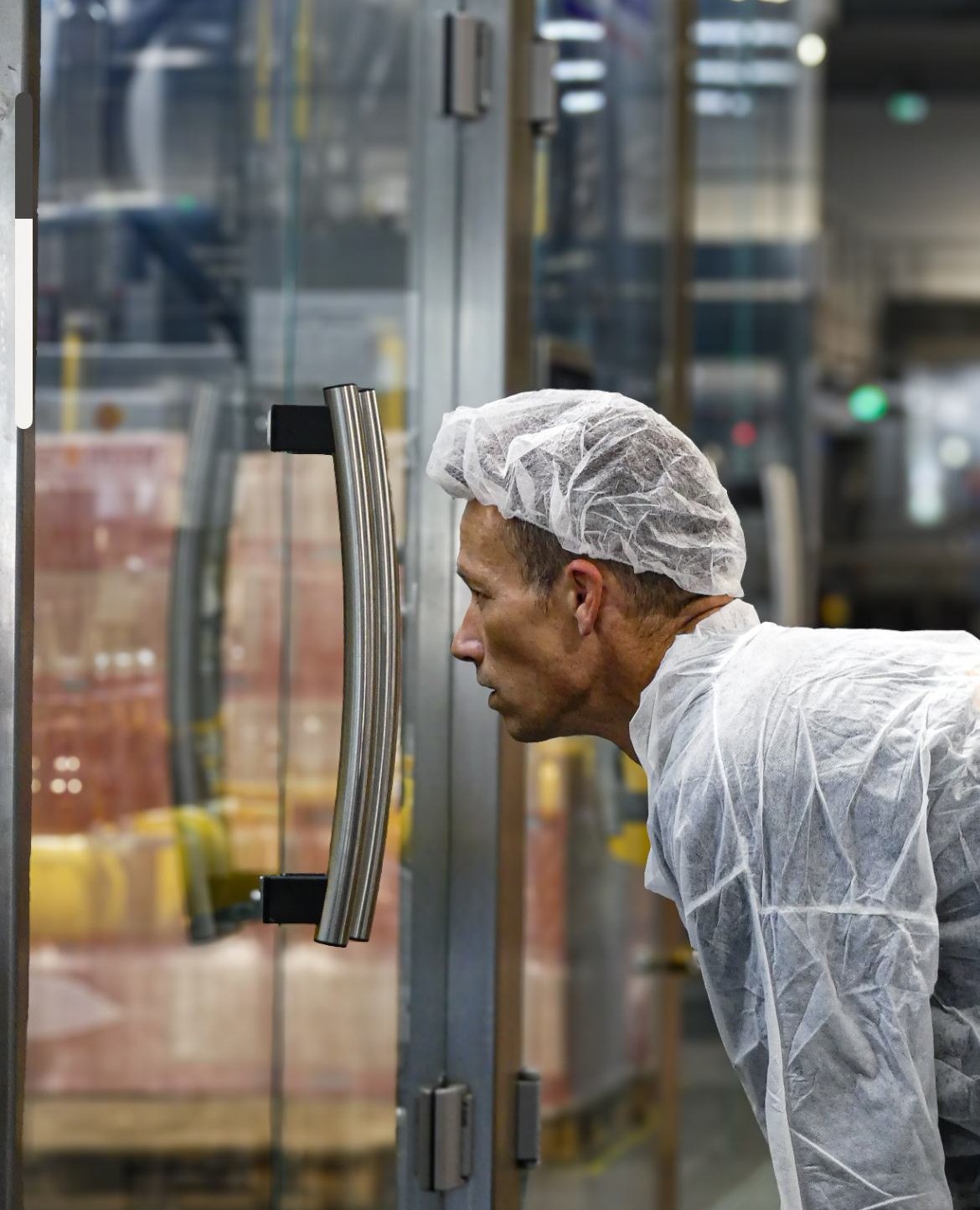 Un opérateur en charlotte et blouse jetable, penché, le visage de profil, qui observe attentivement à travers une porte vitrée. Derrière la vitre, des palettes et la zone de production. Le regard est concentré.